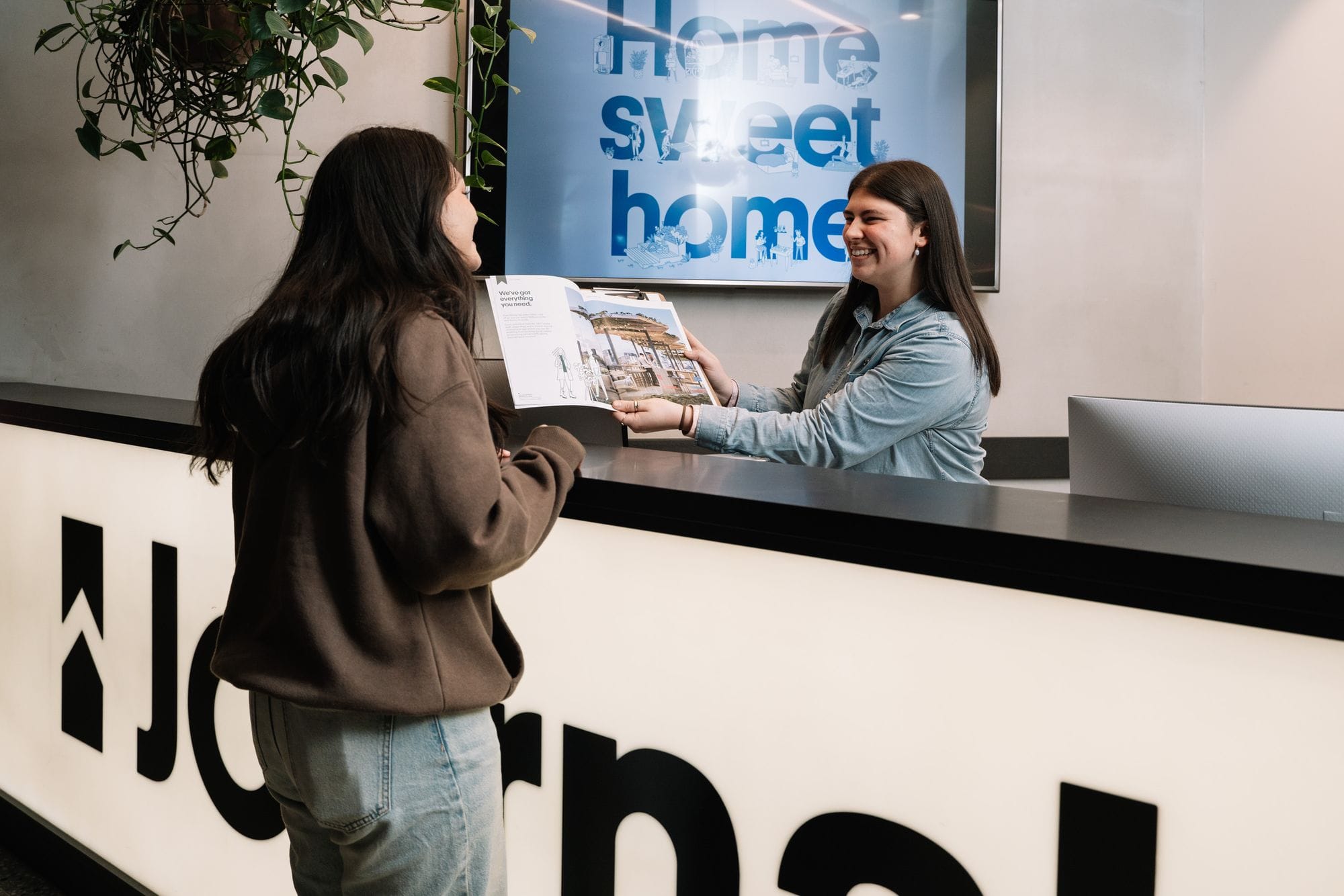 Two people interacting at a reception desk with a “Home sweet home” sign in the background.