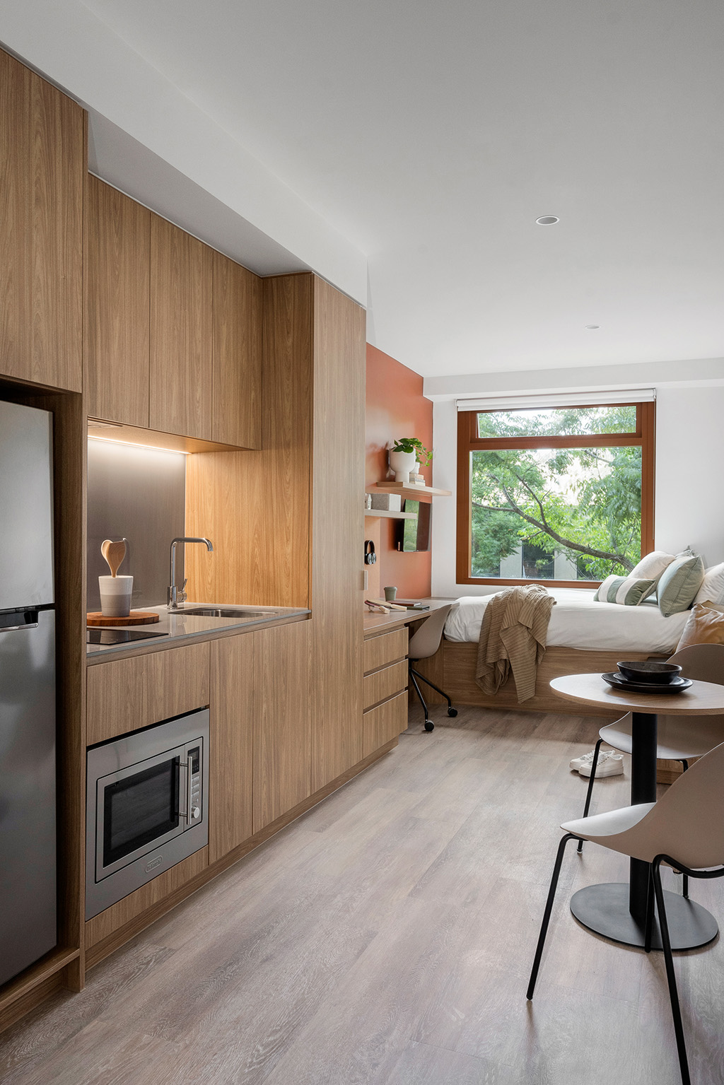 Compact studio with kitchenette, workspace, and bed by a large window.