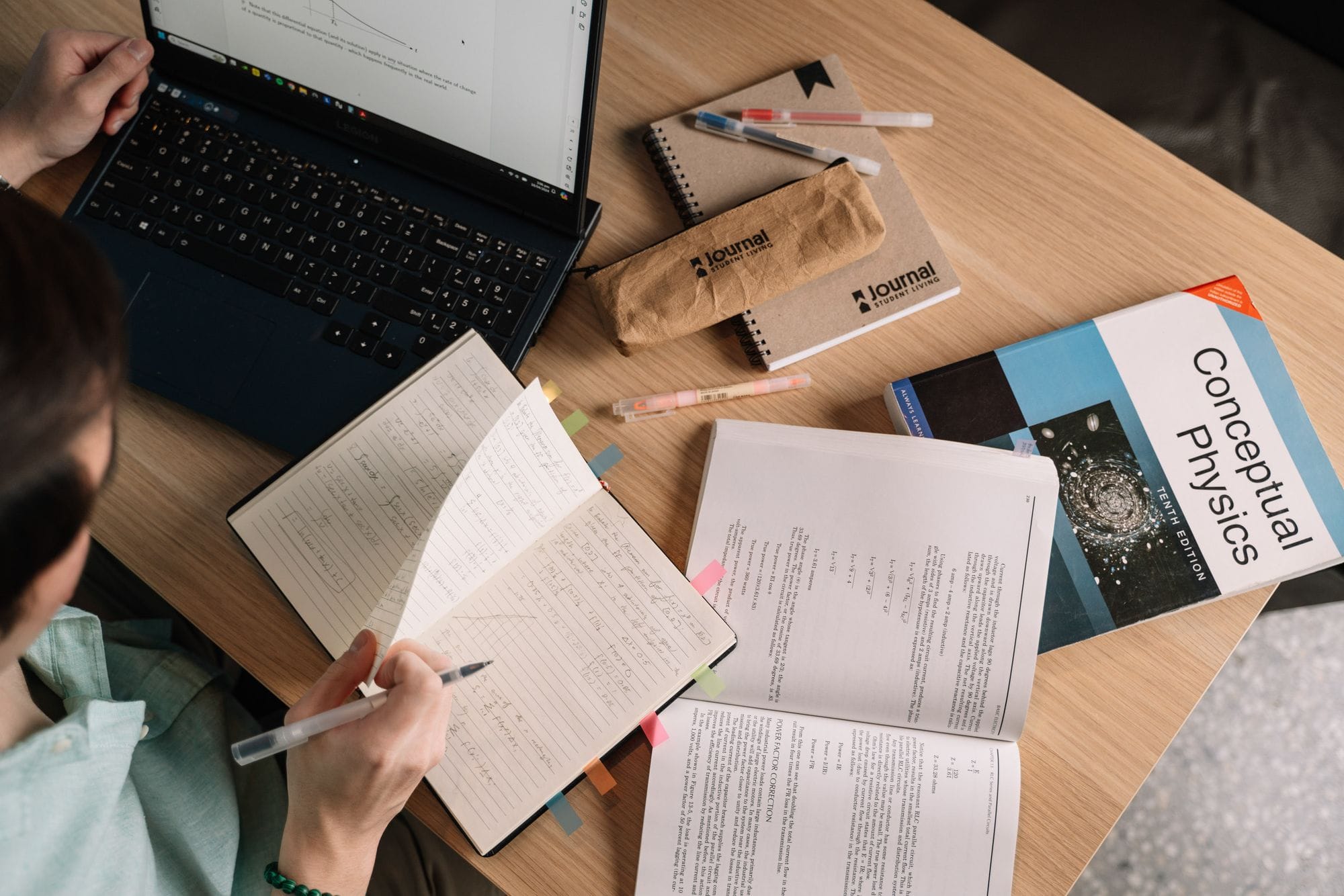 Student writing in notebook at a desk with laptop and study materials.