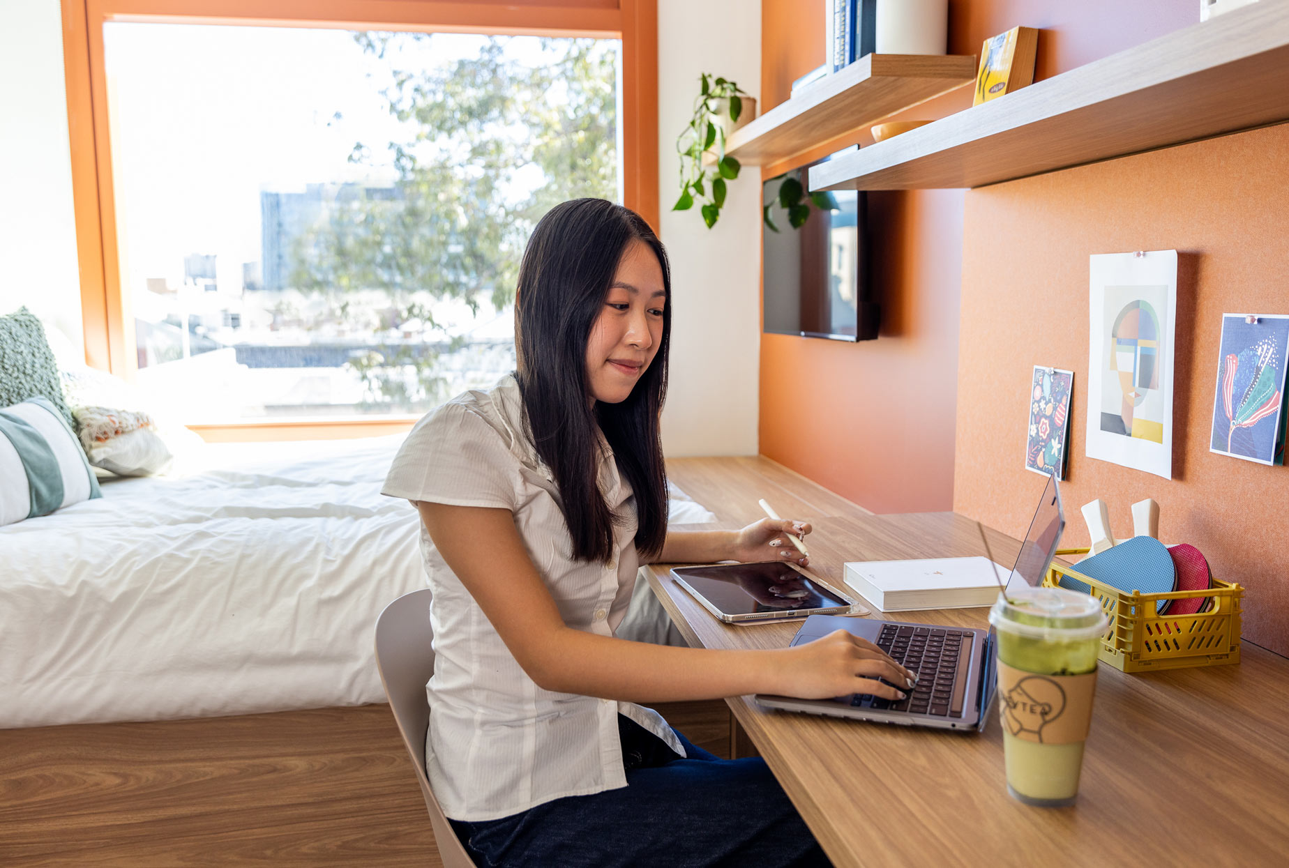 Person working on a laptop at a desk in a modern, well-lit room with shelves and a bed in the background.