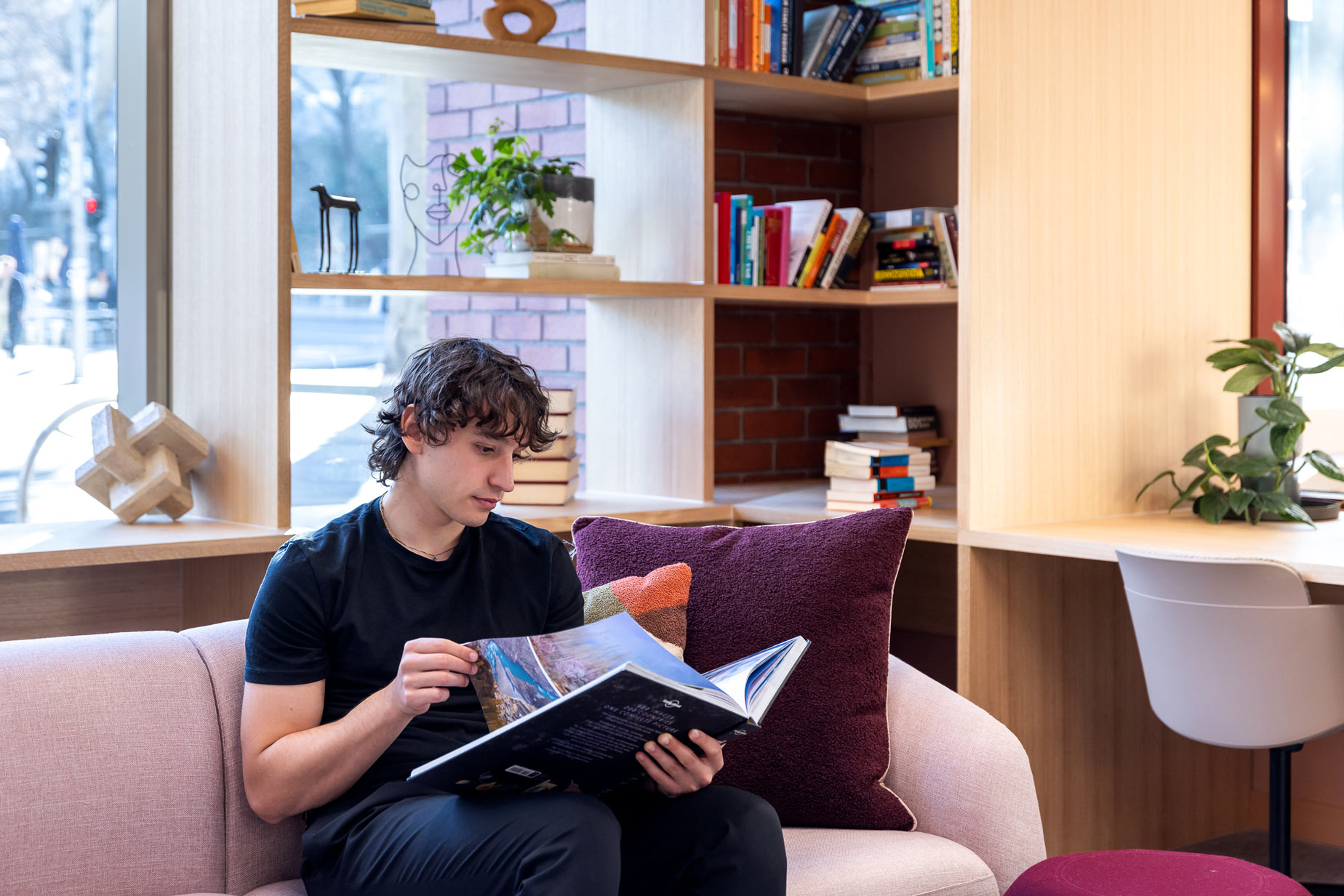 Person reading a large book on a couch in a cosy, sunlit room with shelves and plants.