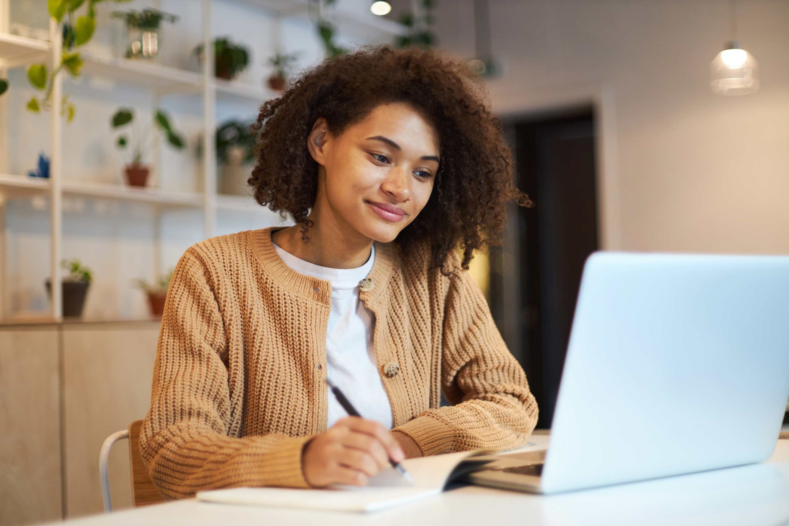Person studying at a desk with laptop, notebook, and indoor plants.